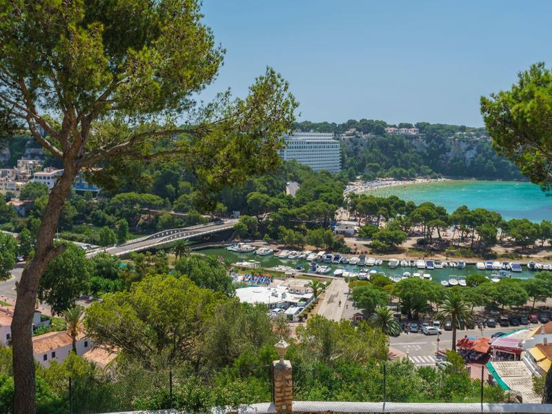 Landschaft mit mediterranen Bäumen, Blick auf einen Strand mit türkisfarbenem Wasser und Ferienhäusern.