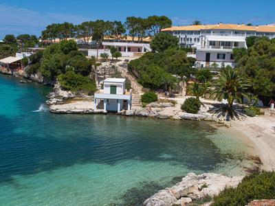 Klares blaues Wasser, Sandstrand und weiße Gebäude mit roten Dächern in grüner Küstenlandschaft.