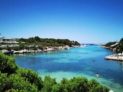 Blaue Lagune mit klarem Wasser, grünen Büschen im Vordergrund und Felsen entlang der Küste.