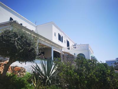 Weißes Gebäude mit Terrasse, umgeben von grünen Pflanzen und klarem blauem Himmel.