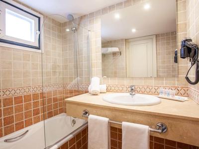 Salle de bain lumineuse avec lavabo, miroir, sèche-cheveux, serviettes et baignoire sous une fenêtre.
