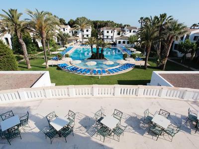 Vista de una terraza de hotel con mesas y sillas, detrás una gran piscina y palmeras.