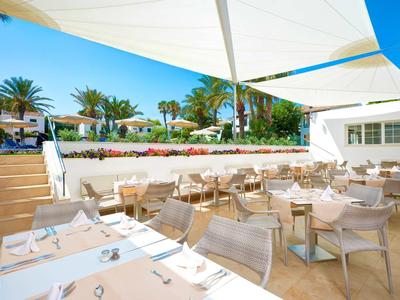 Terrasse de restaurant ensoleillée avec tables et chaises, entourée de palmiers et de toiles d'ombrage bleues.