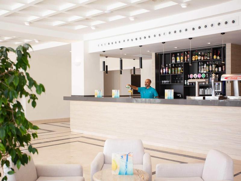 Bar d'hôtel moderne avec comptoir clair, barman et coin salon confortable au premier plan.