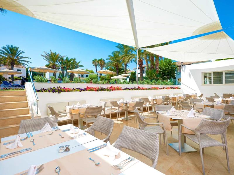 Terrasse de restaurant ensoleillée avec tables et chaises, entourée de palmiers et de toiles d'ombrage bleues.