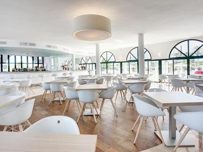 Licht, modern restaurant met witte stoelen, houten vloer en grote bogenramen.