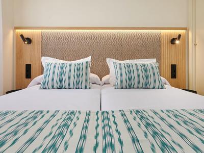 Nowoczesne podwójne łóżko z niebieskimi poduszkami w paski i narzutą pod miękkim zagłówkiem z lampami ściennymi.