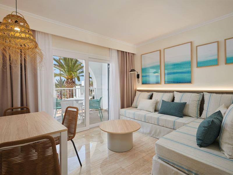 Jasny, nowoczesny salon z wygodnymi sofami, stołem, krzesłami i widokiem na taras z palmami.