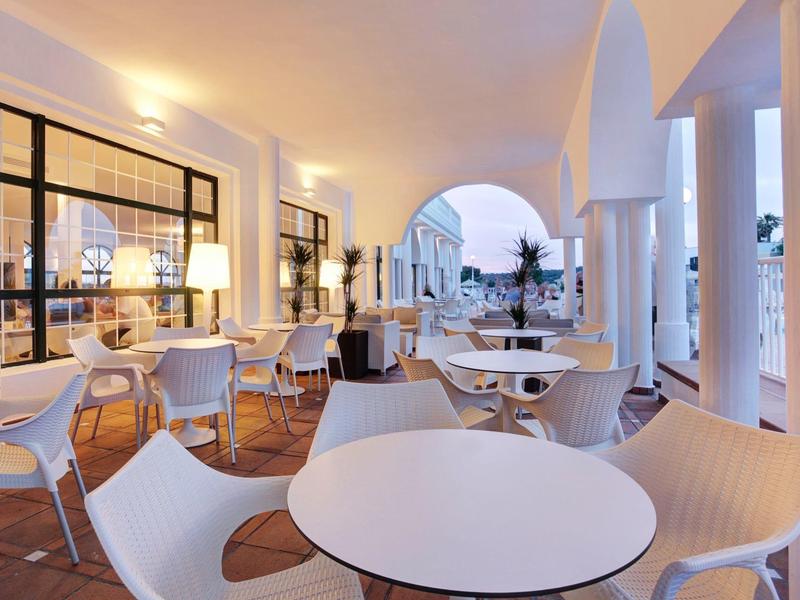 Elegancki taras z białymi stołami i krzesłami pod zadaszonym miejscem z kolumnami.