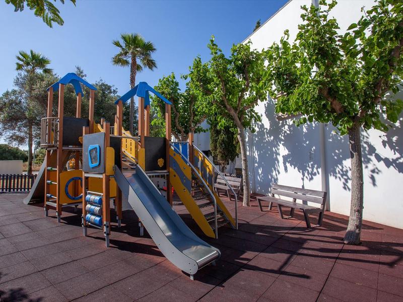 Plac zabaw dla dzieci z zjeżdżalnią i konstrukcją do wspinaczki obok drzew i białej ściany