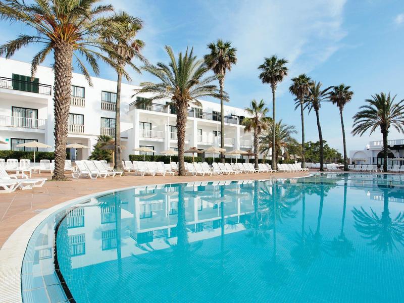 Czysty niebieski basen hotelowy z palmami i białymi leżakami przed białym budynkiem hotelu.