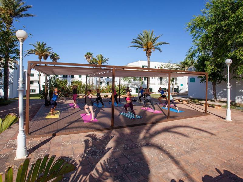 Zadaszona przestrzeń z ławeczkami i ludźmi w słonecznym parku z palmami.