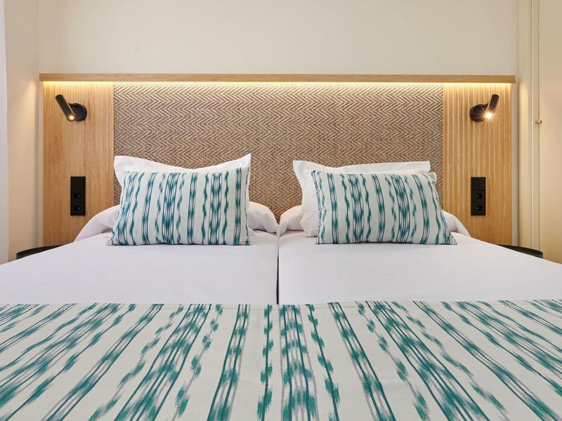 Nowoczesne podwójne łóżko z niebieskimi poduszkami w paski i narzutą pod miękkim zagłówkiem z lampami ściennymi.