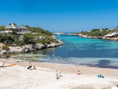 Piaszczysta plaża z małą liczbą osób i czystą niebieską wodą, otoczona skalistymi brzegami i domami.