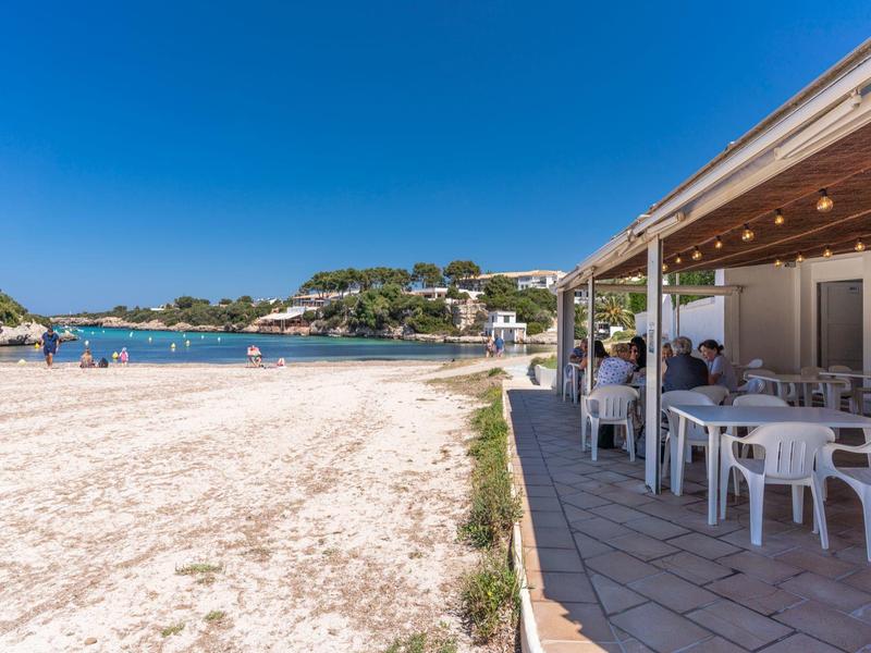 Plaża z białym piaskiem obok otwartej restauracji z białymi stołami i krzesłami pod niebieskim niebem