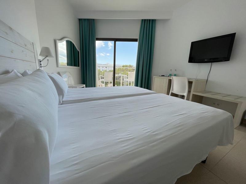 Jasny pokój hotelowy z dużym łóżkiem, zielono-niebieskimi zasłonami i balkonem z widokiem na zewnątrz.