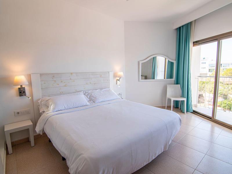 Jasny pokój hotelowy z dużym łóżkiem, stolikami nocnymi i balkonem z widokiem na zieleń.