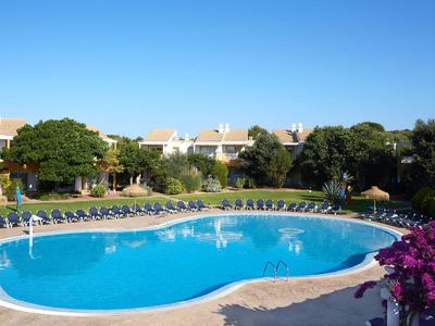 Large pool with many sun loungers next to vacation homes and blooming plants under clear sky.