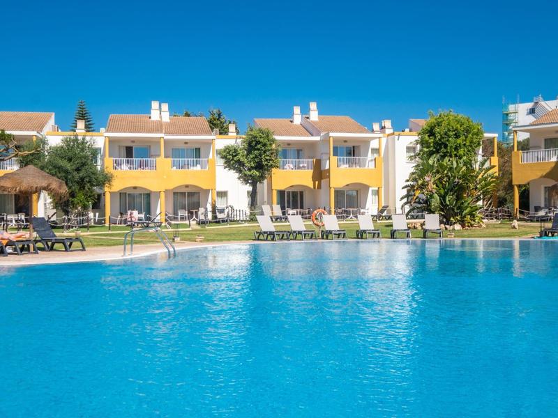 Appartamenti gialli con terrazze accanto a una grande piscina blu sotto un cielo limpido.