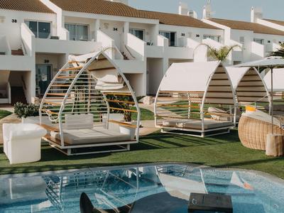 Modern outdoor pool area with lounge chairs and shaded seating in front of white villas.