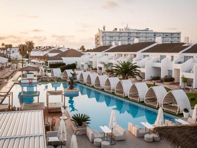 Beach hotel with pool, lounge chairs, and white huts under cloudy sky.