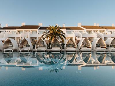 Hotel blanco con ventanas arqueadas y palmeras reflejadas en el agua tranquila de la piscina.