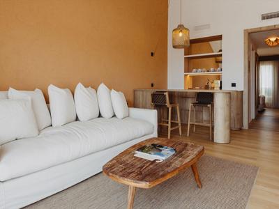 Habitación de hotel luminosa con sofá blanco, mesa de madera y barra en tonos cálidos.
