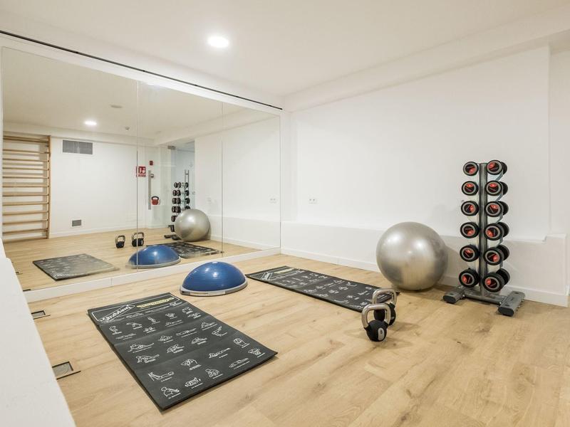 Modern gym room with wooden floor, mirrored wall, exercise mats, and various workout equipment.