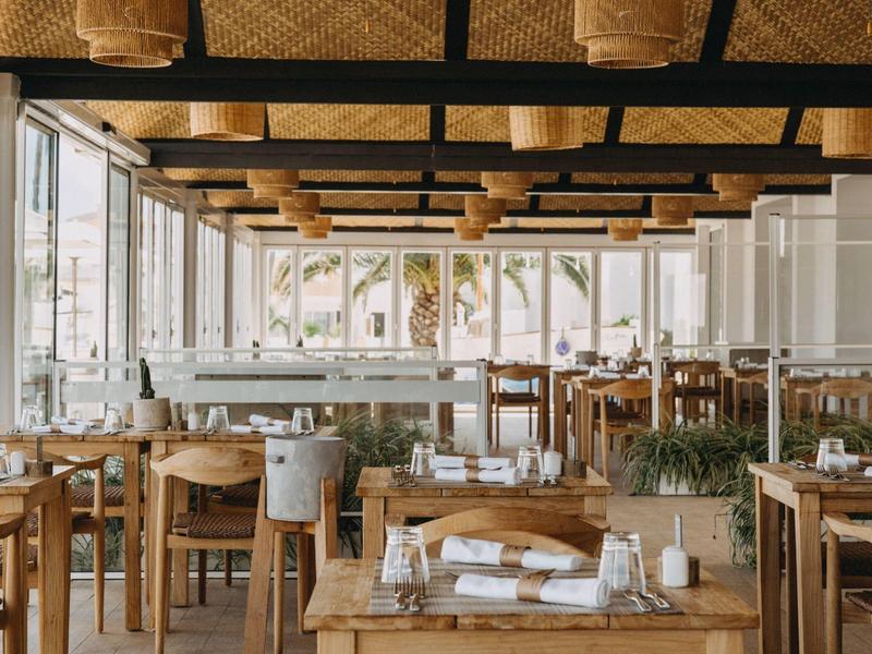 Brightly decorated restaurant with wooden chairs and large windows overlooking palm trees.