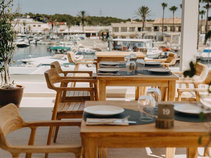 Restaurant terrace with wooden tables overlooking marina and palm trees.