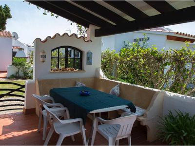 Überdachte Terrasse mit blauem Tisch, weißen Plastikstühlen und bepflanzter Hecke im Hintergrund.