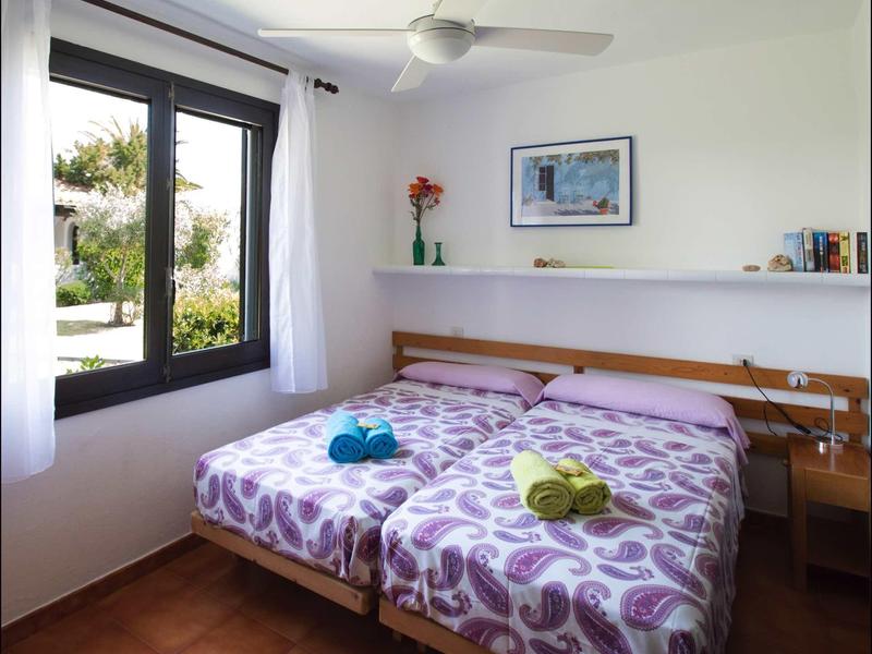 Hell eingerichtetes Schlafzimmer mit zwei Betten, großen Fenster, Ventilator und Wandregal.