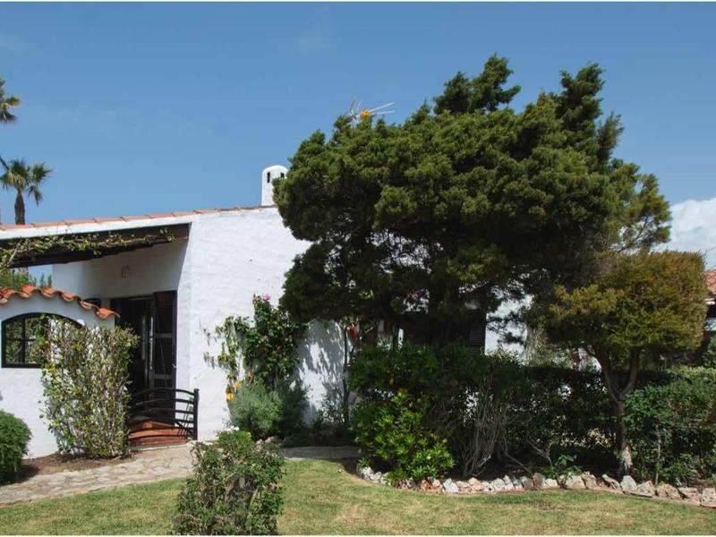 Ein kleines weißes Haus mit braunem Dach, umgeben von grünen Büschen und Bäumen unter blauem Himmel.