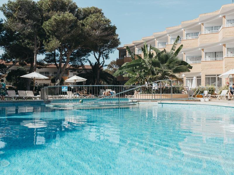 Large hotel pool with sun loungers, umbrellas, and Mediterranean greenery under a clear sky.