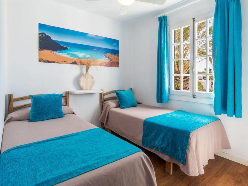 Lichte hotelkamer met twee eenpersoonsbedden, blauwe accenten, een schilderij van een strand en een raam met gordijnen.