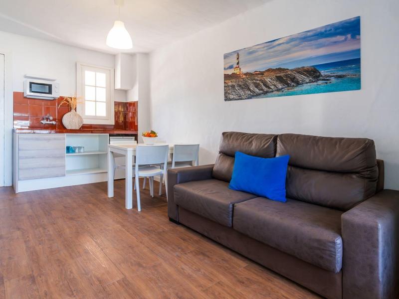 Moderne studio met keuken, bruine bank, blauwe kussen en zeezicht aan de witte muur.