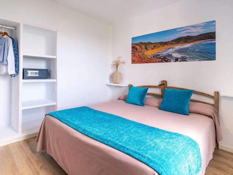 Lichte slaapkamer met tweepersoonsbed, turquoise kussens, wanddecoratie, open kast en houten vloer.