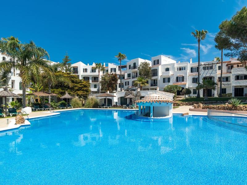Groot buitenzwembad omgeven door witte gebouwen en palmbomen onder een heldere blauwe lucht.