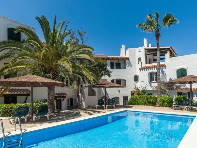 Hotelzwembad met ligstoelen, parasols, palmbomen en witte gebouwen onder een heldere blauwe lucht.