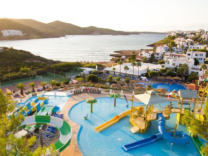 Resort met zwembaden, glijbanen, ligstoelen en uitzicht op zee met heuvels op de achtergrond.