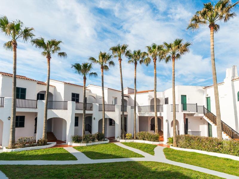 Witte hotelgebouwen met twee verdiepingen en balkons omringen een binnenplaats met palmbomen en groene gazons.