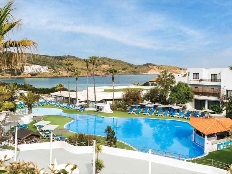 Buitenzwembad van hotel met ligbedden, parasols, palmbomen en zeezicht onder een blauwe hemel.