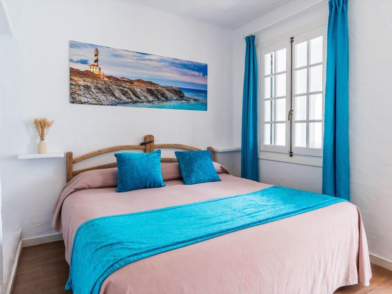 Lichte slaapkamer met tweepersoonsbed, turquoise accenten, groot raam en kustmuurschildering.