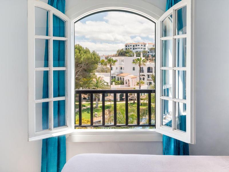 Open raam met blauwe gordijnen met uitzicht op balkon en gebouwen onder bewolkte lucht.