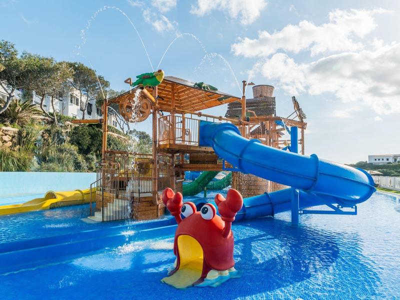 Water speeltuin met glijbanen en waterspelen omgeven door palmbomen en rotskliffen.