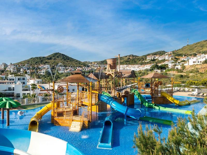 Buitenspeeltuin met meerdere glijbanen en zwembaden omringd door heuvels en gebouwen.