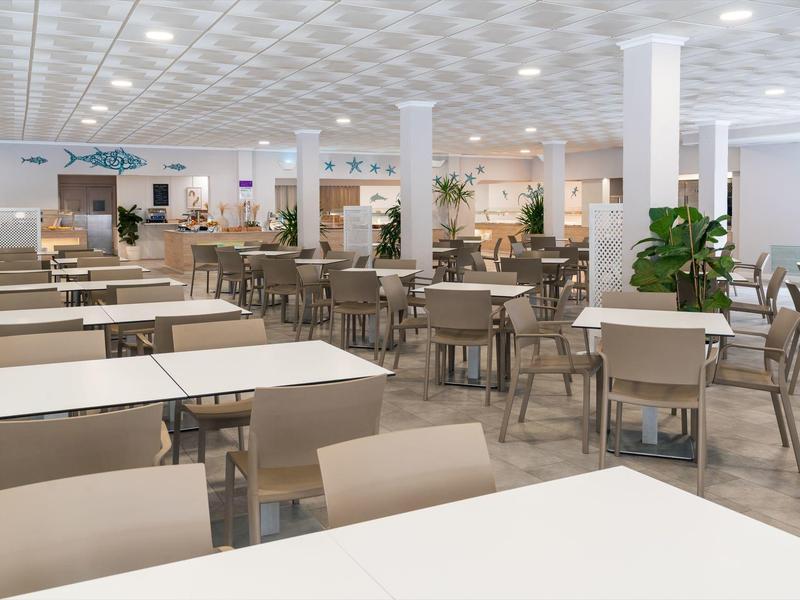 Ruime, moderne eetzaal met witte tafels, veel stoelen en zachte bovenverlichting.