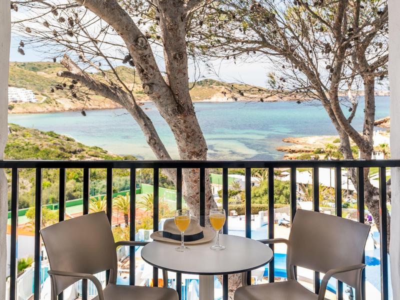 Balkon met twee stoelen en een tafel met uitzicht op een strand met parasols en bomen.