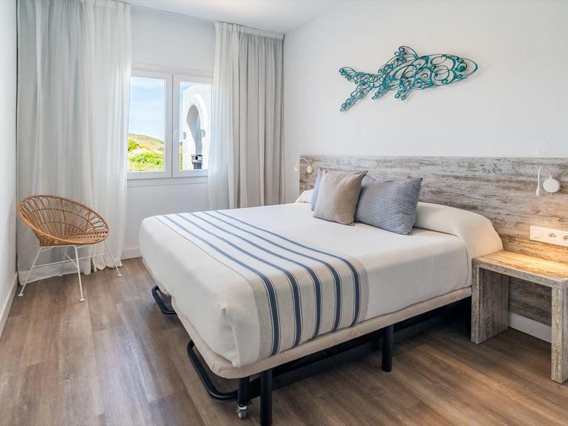 Lichte hotelkamer met tweepersoonsbed, houten vloer, stoel en raam met witte gordijnen.