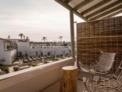 Balcony with wicker chairs overlooks a modern hotel pool area with palm trees under cloudy sky.